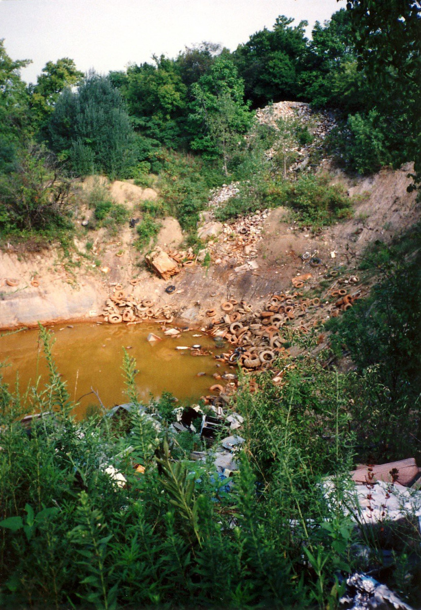 A small body of orange-hued water surrounded by waste in a wooded area.