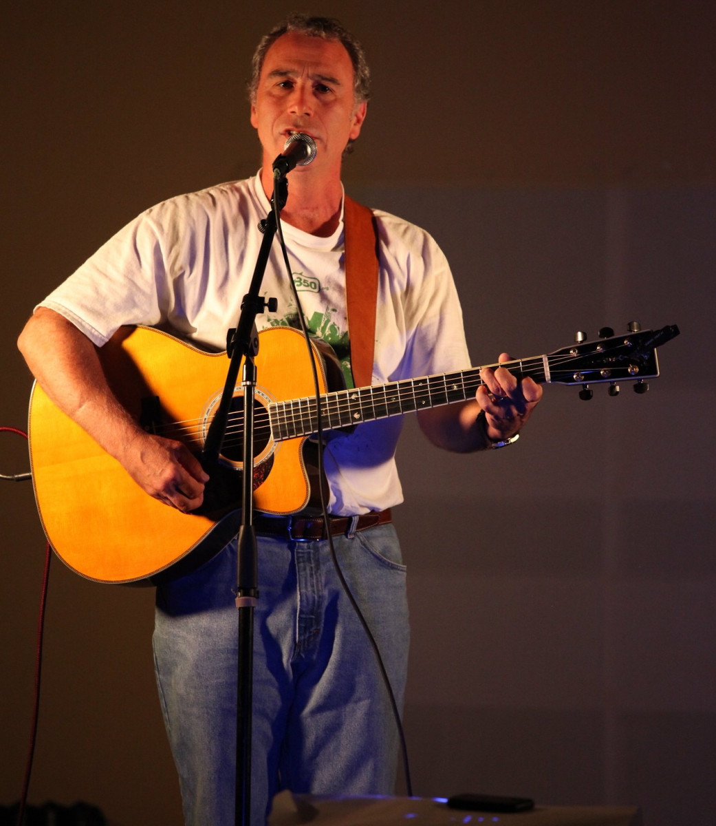 Joe Uehlein sings into a microphone while strumming an acoustic guitar.