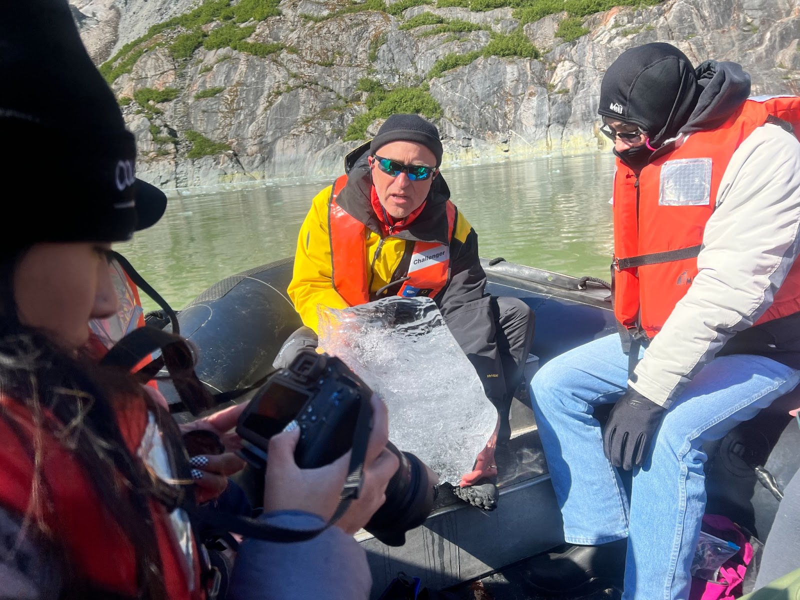 A white male in a knitted hat, wearing a bright orange life vest, holds a large piece of ice he plucked from the water. He speaks to other people aboard a small boat, also wearing warm gear and orange life vests.