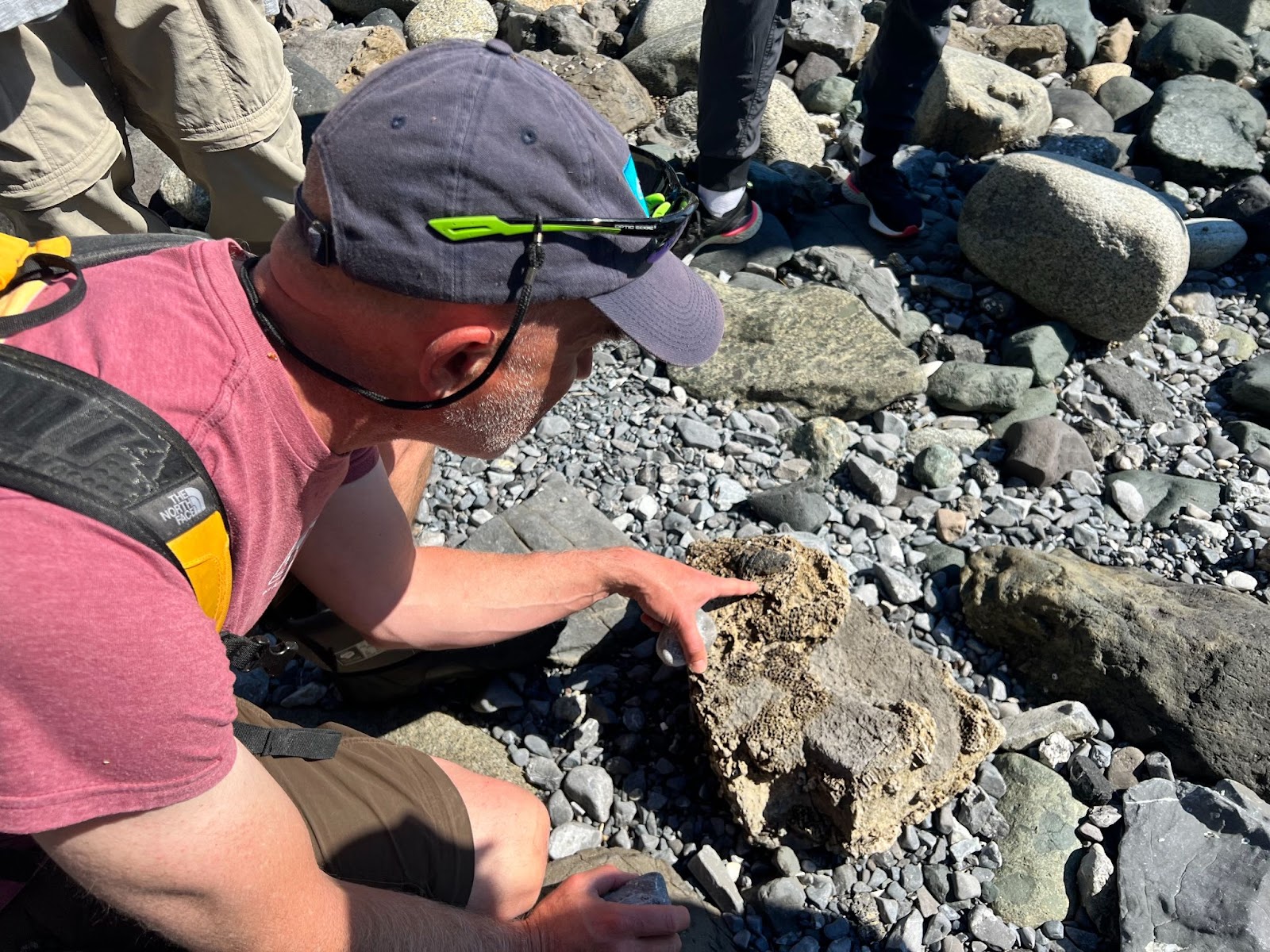 A man in a red shirt and wearing a grey baseball cap points at a rock amid a field fo grey rocks and small boulders.